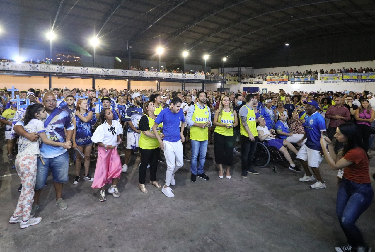 Na foto, a secretária Silvia Grecco segura o microfone e discursa ao público. Ao lado esquerda dela está o presidente da Liga-SP e da Águia de Ouro, Sidnei Carriuolo e Vanessa Dias, da escola Rosas de Ouro. Ao lado direto, está o prefeito Ricardo Nunes e seu filho Nickollas Grecco. Ao fundo, há integrantes da escola e a bandeira da Águia de Ouro, todos estão no palco.